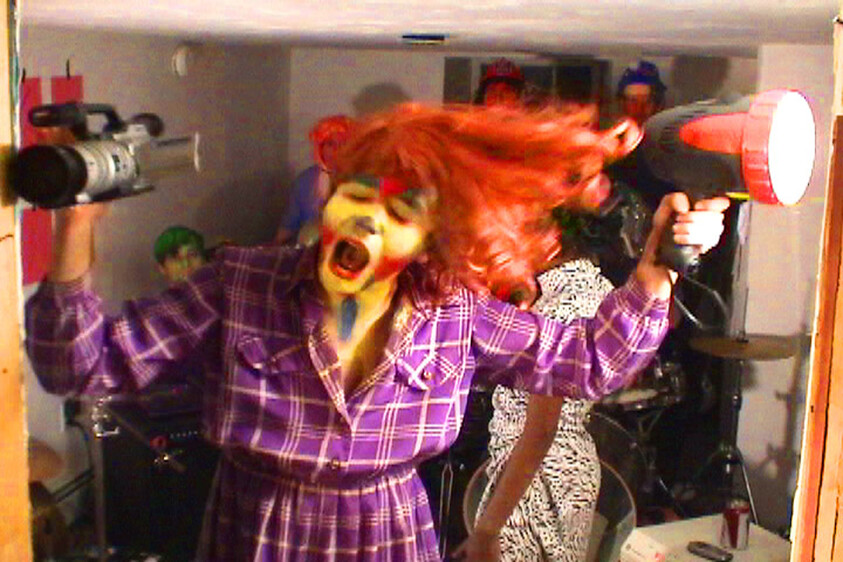 Person in a purple plaid dress with colorful face paint, holding a megaphone and camera, hair flying, in a lively room with instruments.