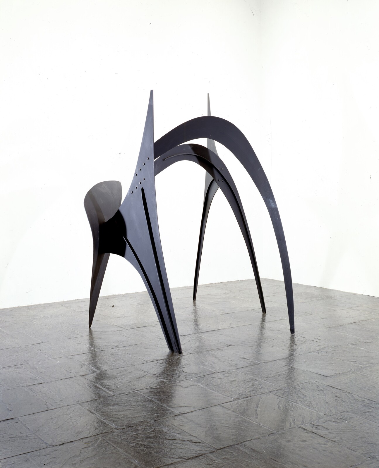 Black metal abstract sculpture with tall curved legs standing on a tiled gallery floor.