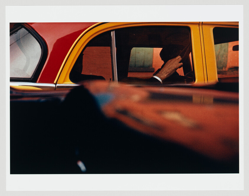 Driver's hand on the steering wheel inside a yellow and red car seen through the window.
