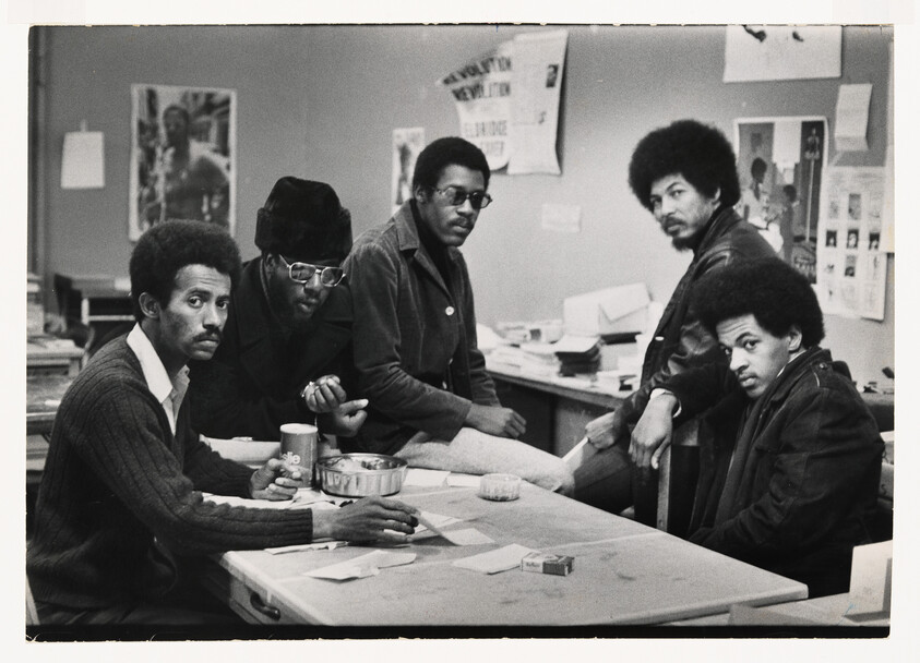 Five men gathered around a table in a room with activist posters, looking intently at the camera.