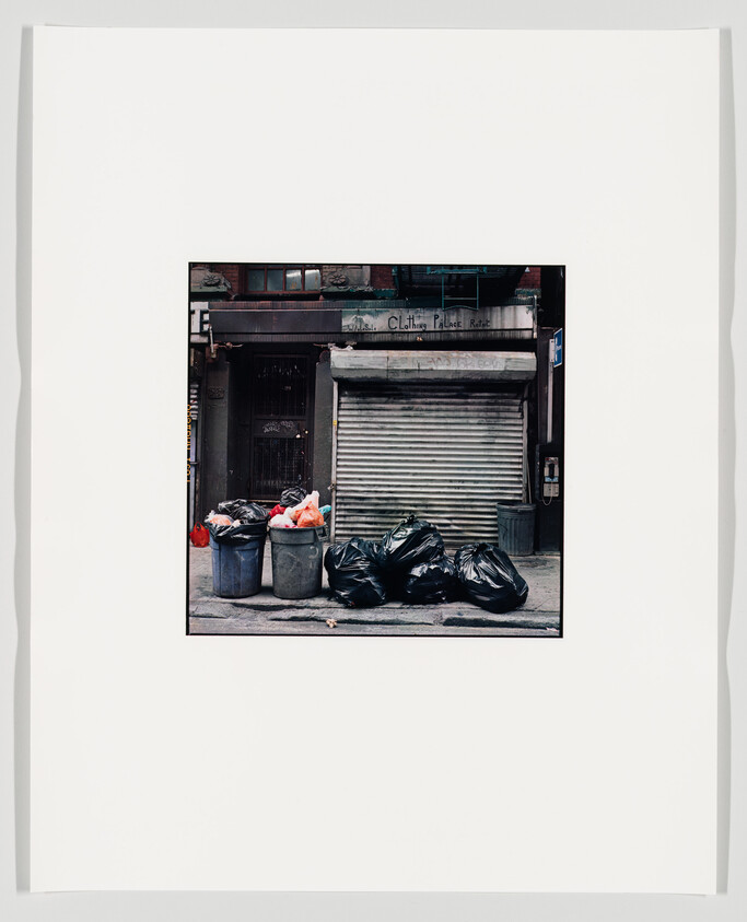 A photograph of a closed storefront with a metal shutter, flanked by trash cans and black garbage bags on the sidewalk. The image is mounted on a white background with a wide border.