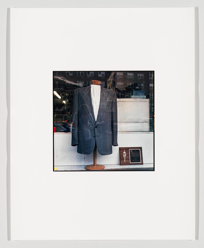 A framed photograph of a mannequin torso dressed in a tailored suit and tie displayed in a storefront window, with reflections of the street visible on the glass.