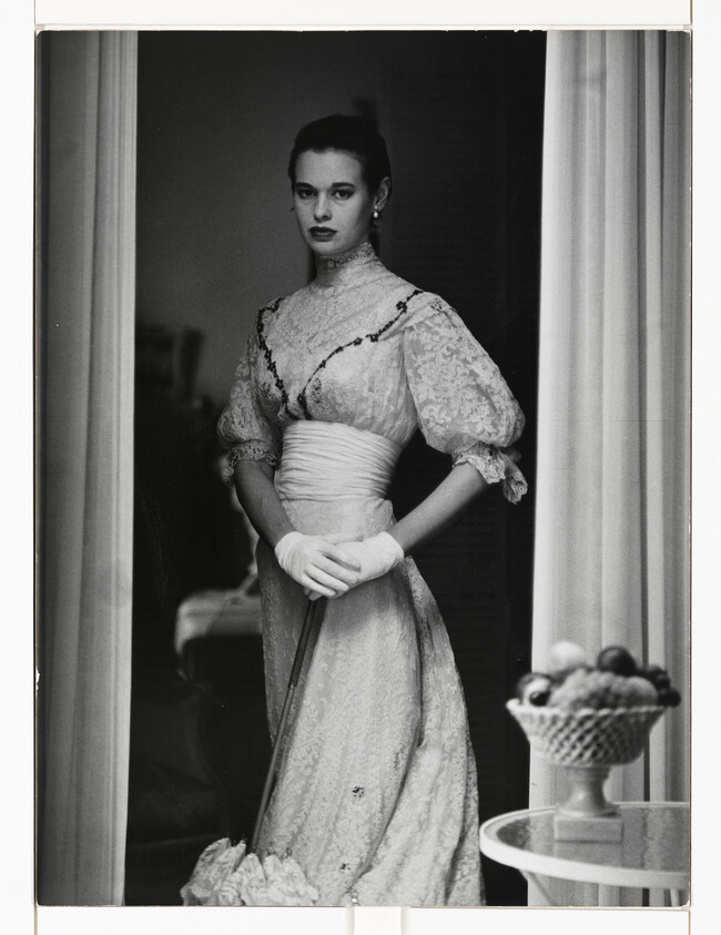 Young woman in a high-necked lace dress and gloves stands holding a folded parasol.