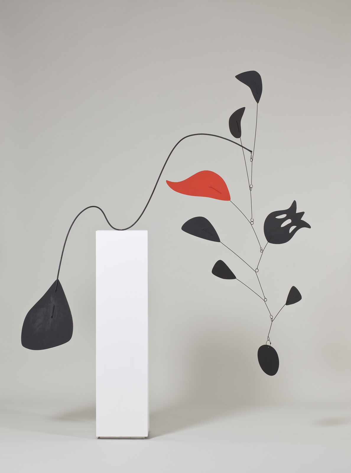 A balanced mobile sculpture on a white pedestal with black shapes and one red leaf.