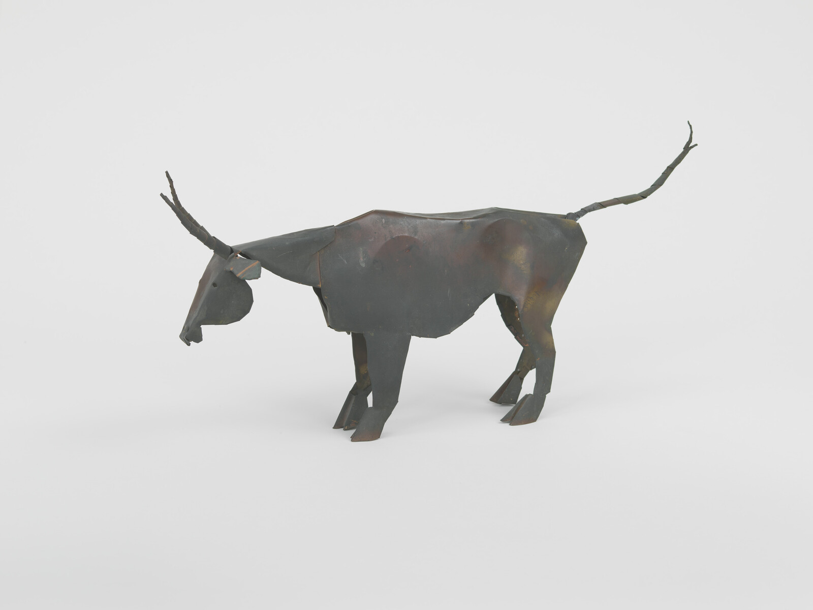 Rusty metal sculpture of a horned cow made from folded sheets standing on a white floor.