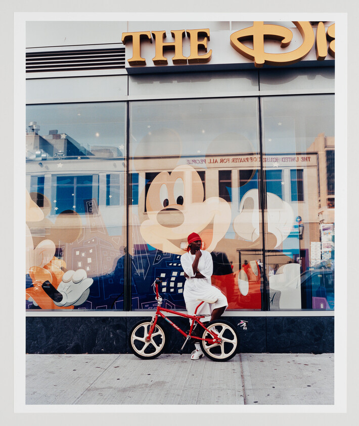 Man in white clothes sits on a red bicycle in front of a store window with giant Mickey Mouse.