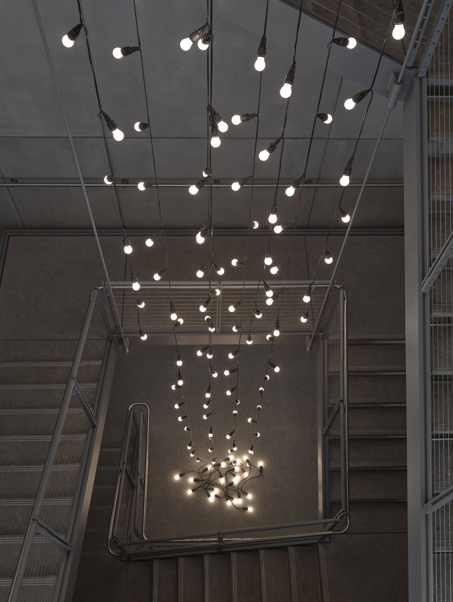 A cluster of hanging light bulbs with glowing filaments suspended from the ceiling by black cables, creating a dynamic lighting installation above a staircase in a room with industrial-style concrete walls and metal railings.