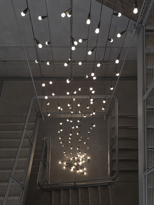 Strings of hanging light bulbs cascade down a stairwell, with several bulbs coiled on the floor.