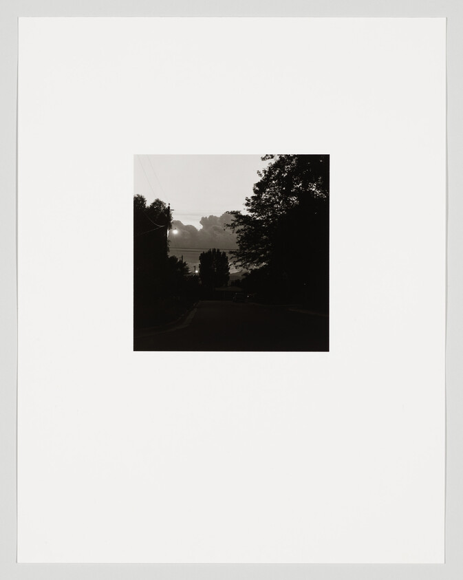 A dark neighborhood street framed by trees with the sun breaking through clouds in the distance.