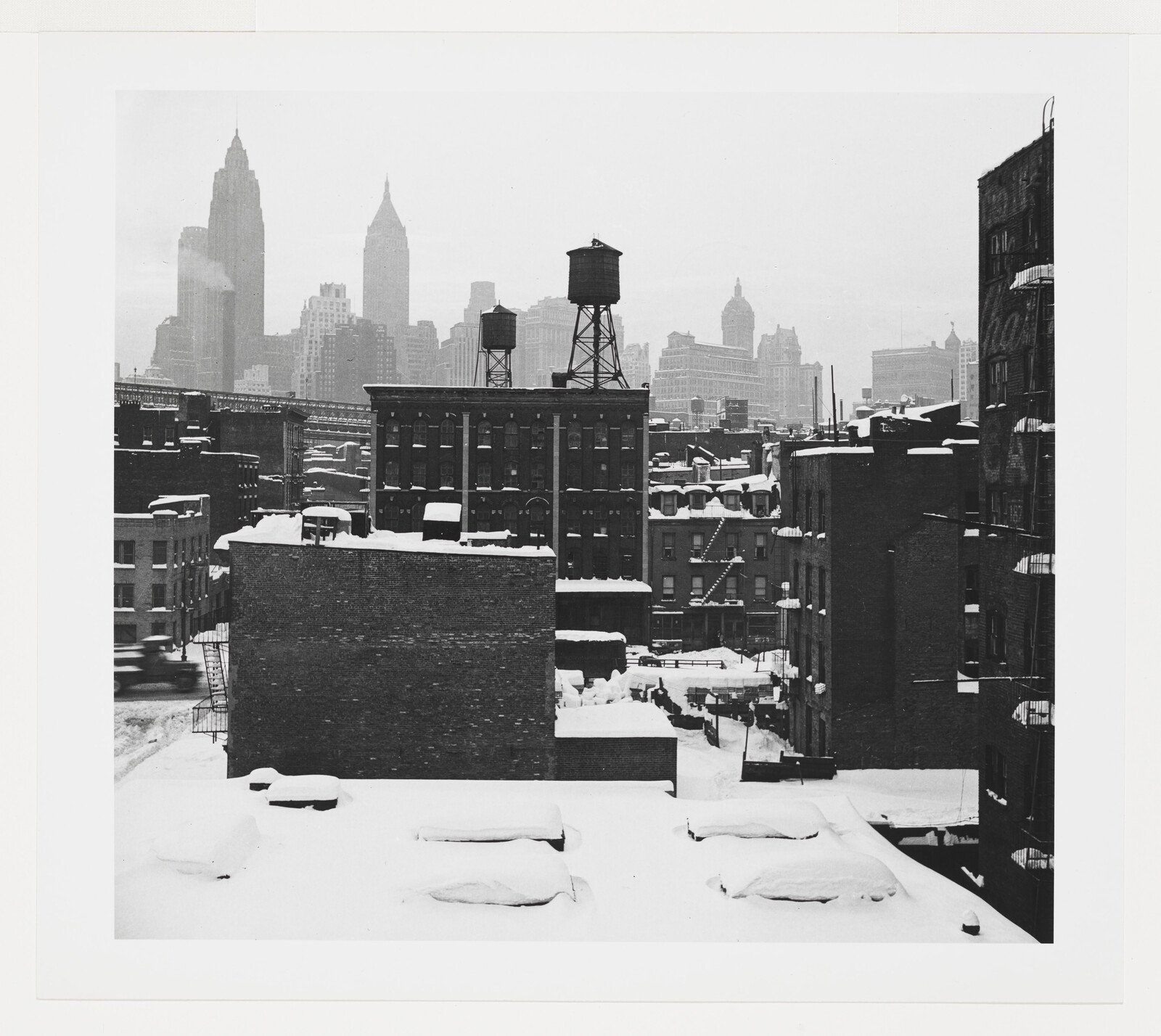Snow-covered rooftops and two rooftop water towers overlooking a distant city skyline.