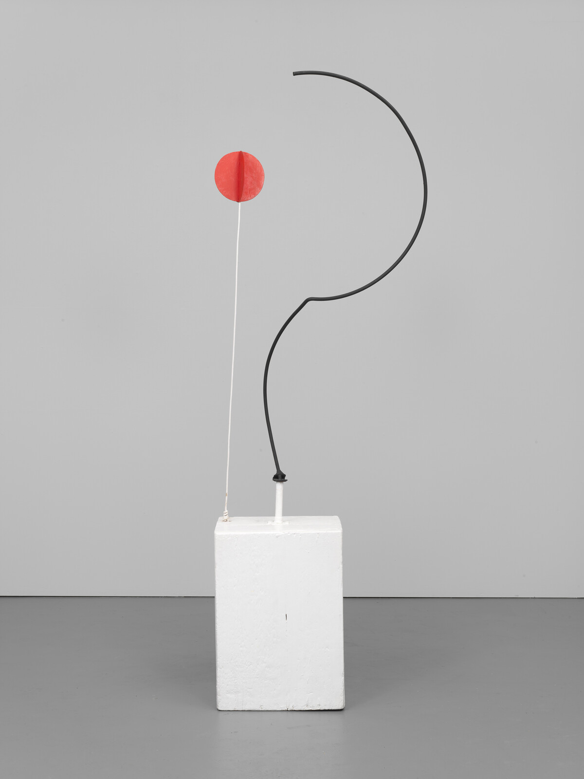 Minimal sculpture with a white pedestal supporting a black curved rod and a thin rod topped by a red disk.