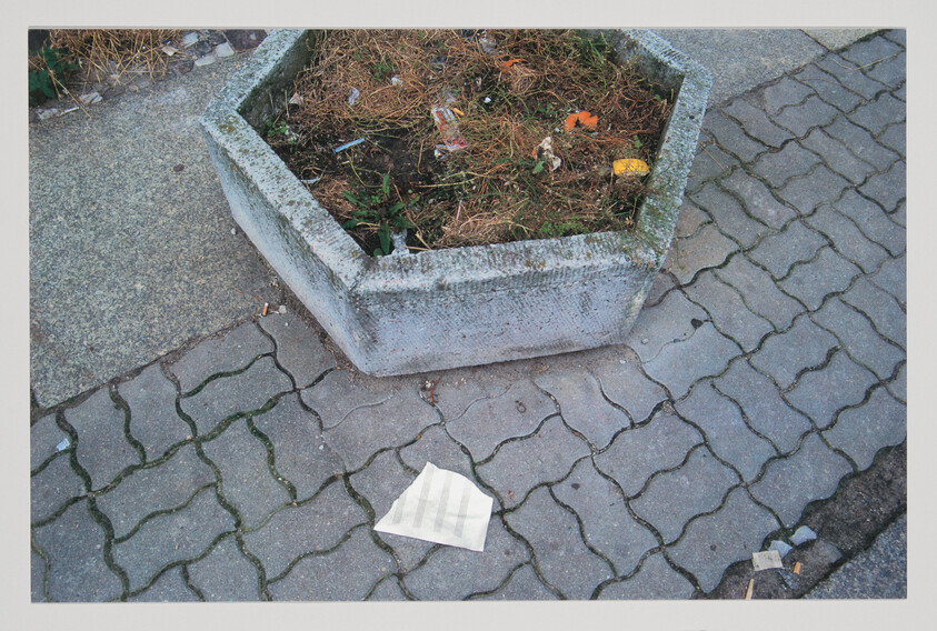 Concrete hexagonal planter with dry weeds and litter beside a discarded white paper on the sidewalk.
