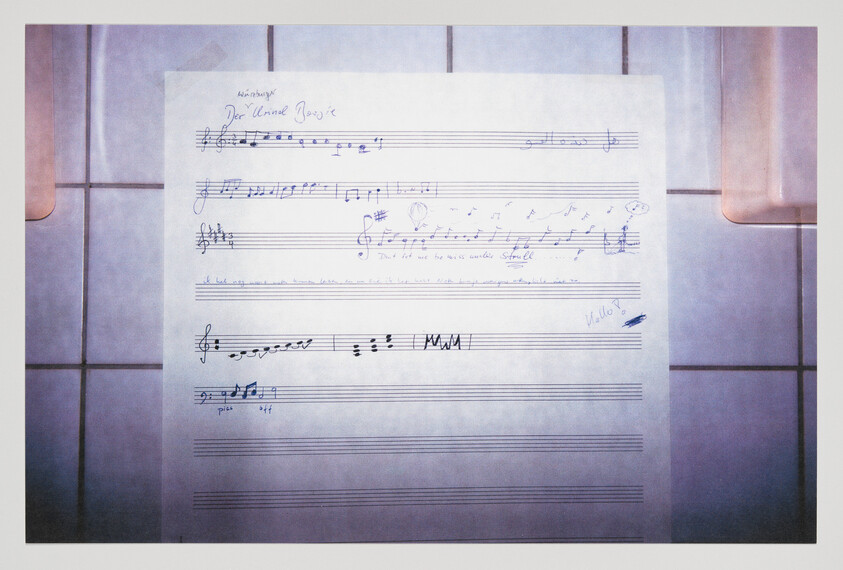 A sheet of music paper with handwritten musical notes and annotations is placed on a tiled surface. The top of the sheet features the title "Der Lindenbaum" in cursive script, and there are various musical notations and dynamic markings throughout the staves. The bottom staves are blank, and there is a signature at the bottom right corner. The lighting suggests an indoor setting with a reflective surface to the right.