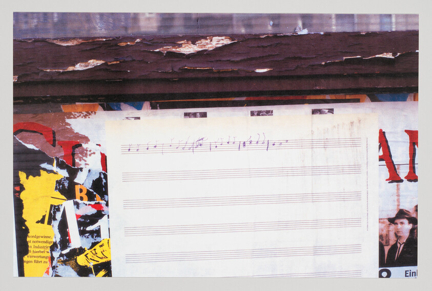 Blank sheet music with a short handwritten musical phrase taped on a torn poster-covered wall.