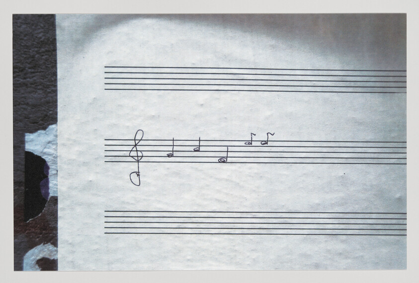 A close-up photo of a piece of paper with musical notation, showing a treble clef and a few notes on the staff, with a slightly crumpled texture and the edge of another object partially visible on the left side.