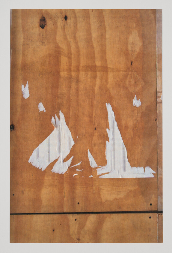 Worn plywood panel with torn white paper remnants forming abstract sail-like shapes.