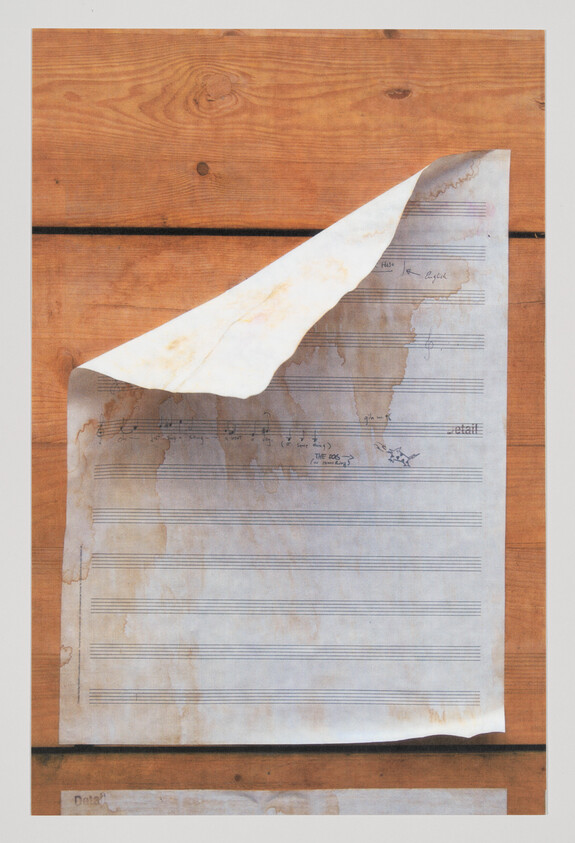 A weathered piece of sheet music with faded notes lies partially unfolded on a wooden surface, showing signs of age and wear with its yellowed edges and coffee-stained appearance.
