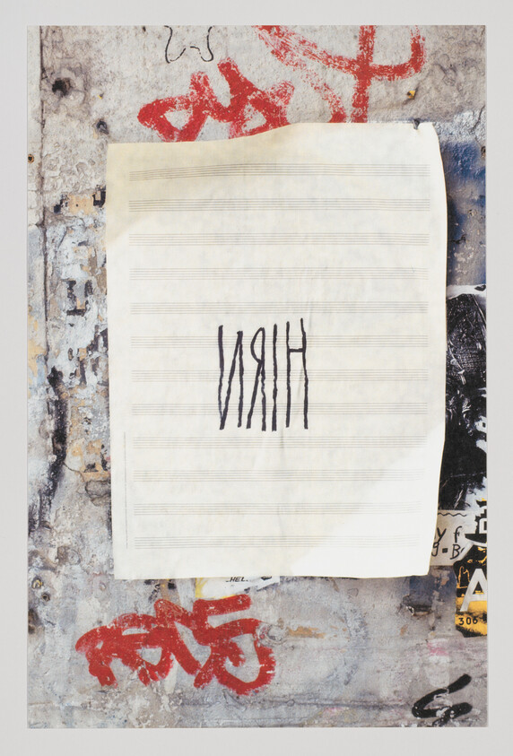 A blank sheet of music paper taped to a graffiti-covered wall with hand-drawn vertical letters.