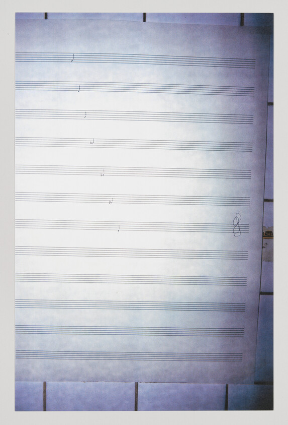 A photograph of a blank sheet of music paper with a few scattered notes written on it, displayed against a blue-tinted background. The treble clef is visible on the right side of the sheet.