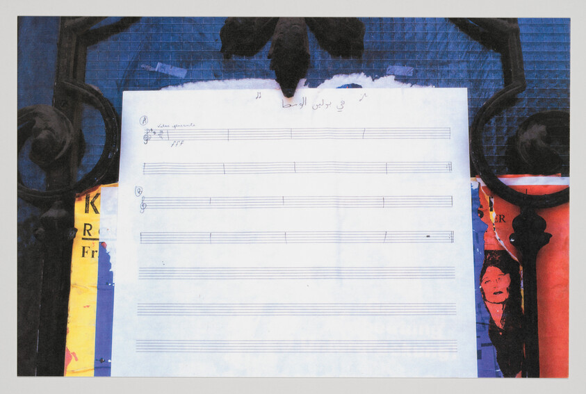 Blank sheet music paper pinned to a decorative iron gate with colorful posters behind.