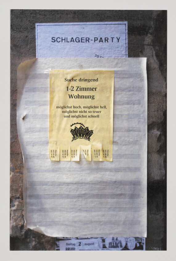 A photograph of a bulletin board with multiple layers of flyers, the topmost being a yellow flyer in German seeking urgently a 1-2 room apartment, with tear-off contact tabs at the bottom. Behind it, partially visible, is a blue flyer for a 'Schlager-Party' and another flyer with musical notation lines. The board appears weathered, and the flyers show signs of wear and tear.