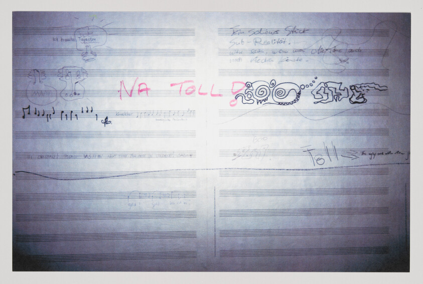 A photograph of a piece of paper with musical staff lines and various doodles and writings in both pen and highlighter. The central doodle is the word "NA TOLL" written in large, pink highlighter letters. Other doodles include a small figure saying "ich brauche Tagesform," musical notes, and a sketch of a fantastical creature. Handwritten text and scribbles are scattered throughout the page.