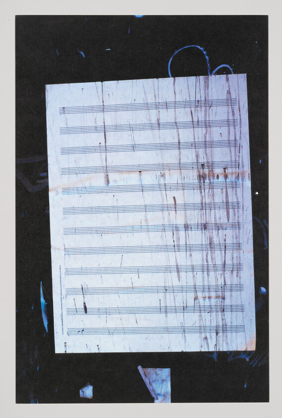 A worn and stained piece of sheet music with empty staves is centered against a dark background with scattered blue paint splatters.