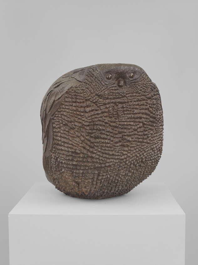 A rounded carved wooden owl sculpture with textured feather details sitting on a white pedestal.