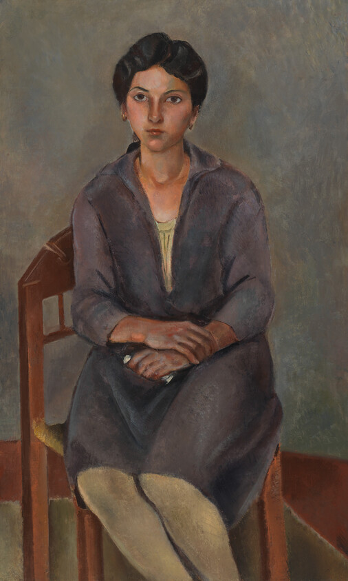 Young woman sits on a wooden chair with hands folded in her lap, looking forward.
