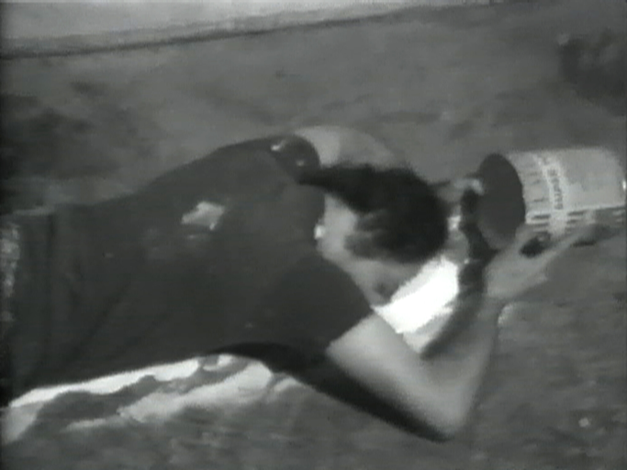 Person lying on the ground, pouring white paint from a large can onto the surface in front of them.
