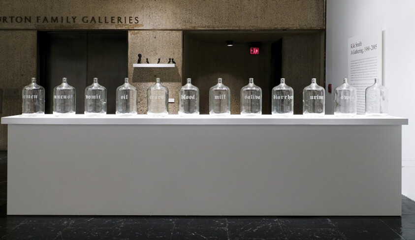 A row of labeled glass jars on a white gallery plinth listing bodily substances like blood and saliva.