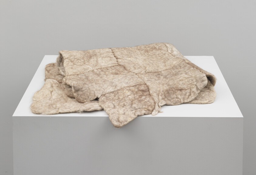 A folded beige felted wool blanket with stitched squares rests on a white display pedestal.