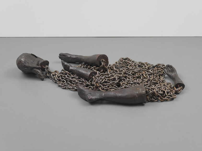 Dismembered bronze limbs connected by heavy chains lie piled on a gray gallery floor.