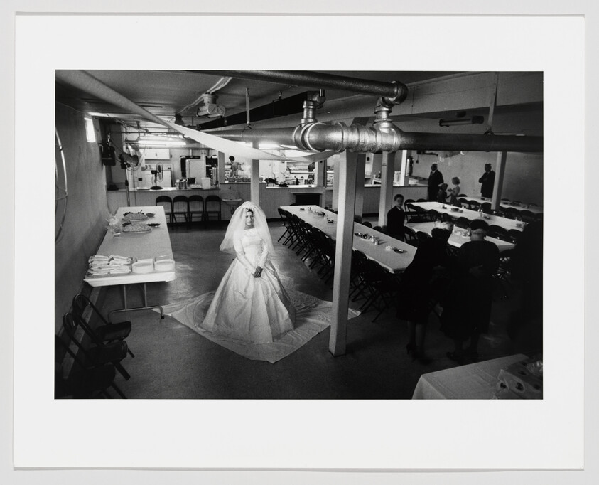 A black and white photograph capturing a bride in a full wedding dress standing alone in the center of a sparsely decorated basement room with exposed pipes overhead. Tables are set up for a reception with guests scattered around, some seated and some standing in small groups.