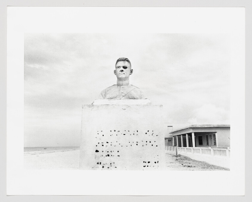 A black and white photograph depicting a bust of a man with a stern expression, mounted on a concrete pedestal with small holes, set against a cloudy sky. The scene is on a beach with an empty horizon and a single-story building with a colonnade to the right.