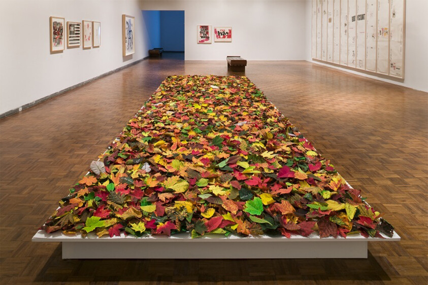 A long rectangular platform covered in colorful autumn leaves stretches through a quiet gallery room.
