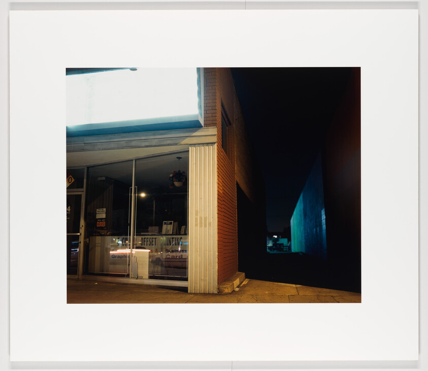 Empty printing shop front with glass doors lit at night beside a dark alleyway.