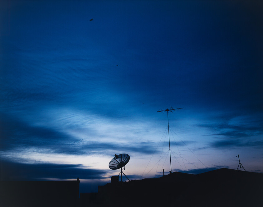 Satellite dish and tall antenna silhouette on a rooftop against a deep blue dusk sky.