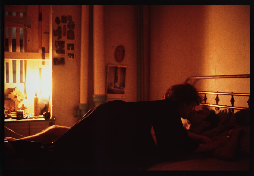 Two people share a kiss while lying on a dimly lit bed in a cozy bedroom.