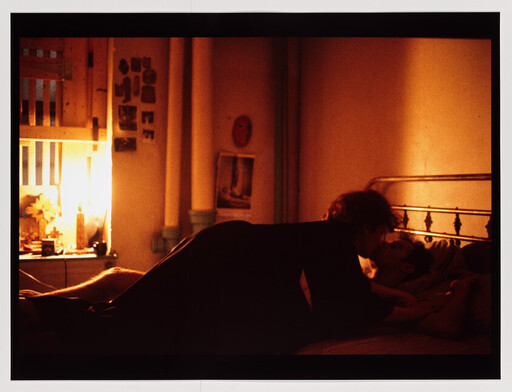 Two people kissing on a bed in a dim, warm-lit bedroom.