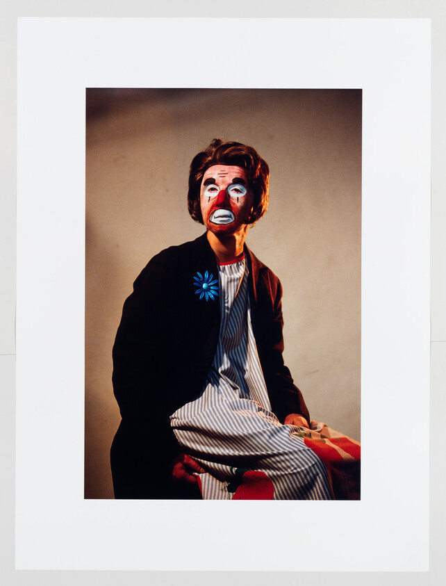 A seated person wearing clown makeup and a dark jacket with a bright blue flower brooch.