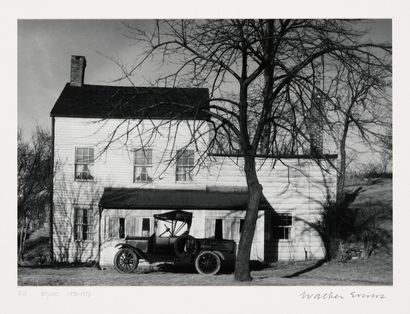 A vintage open-top car parked in front of a two-story white house with a leafless tree.