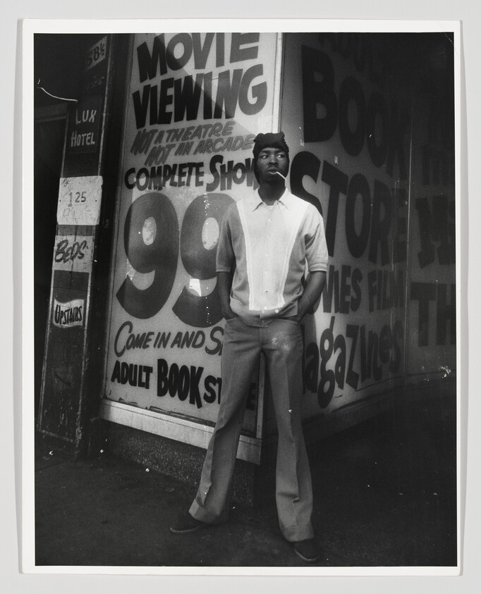 A man stands with hands in pockets in front of a storefront advertising 99-cent movie viewing and adult bookstore.