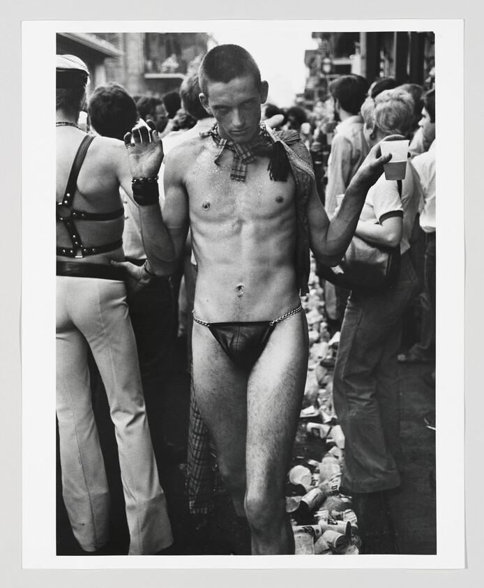 A shirtless man in a leather thong walks through a crowded street holding a cup.