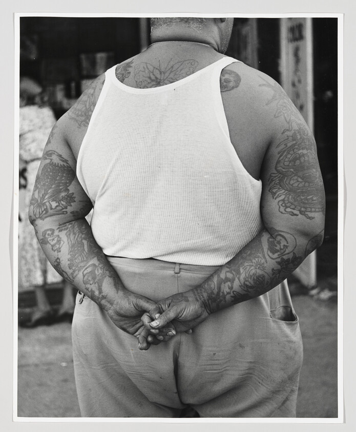 A tattooed person in a white tank top stands with hands clasped behind their back.