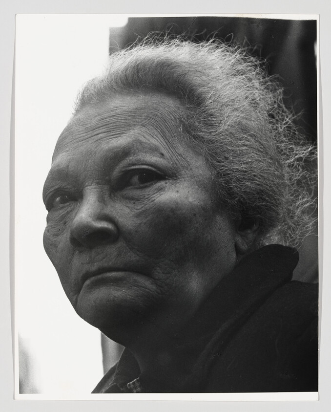 Elderly woman with gray hair looks intently toward the camera with a serious expression.