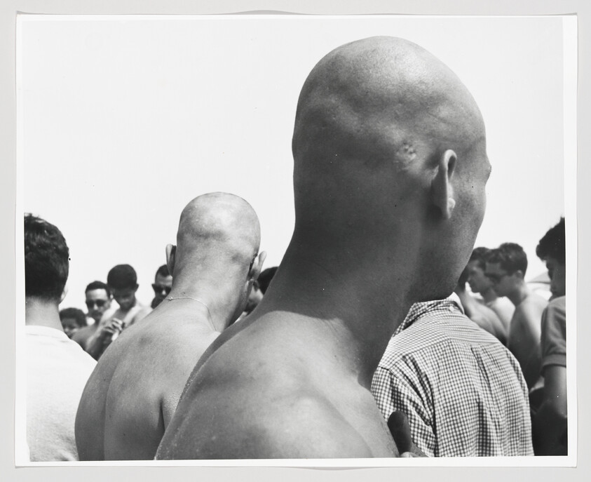 Two shirtless men with shaved heads stand among a crowd of onlookers at an outdoor gathering.