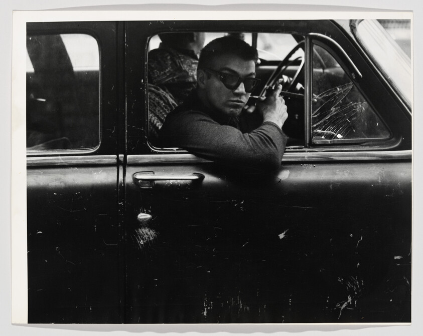Man wearing sunglasses leans out of an old car window holding a cigarette and looking back.