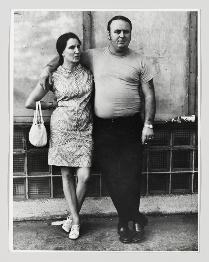 A man stands with his arm around a woman as they lean against a wall.