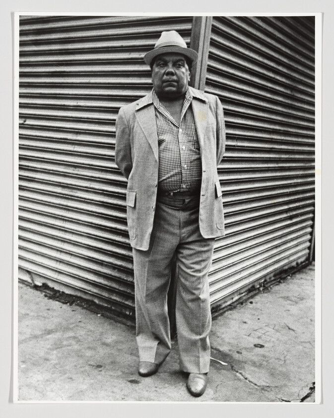 A man in a suit and hat stands with hands behind his back against a corrugated metal shutter.